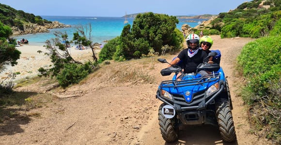 Three blue quad bikes and riders in helmets on a dirt viewpoint above green hills, with a bay and headland in the distance