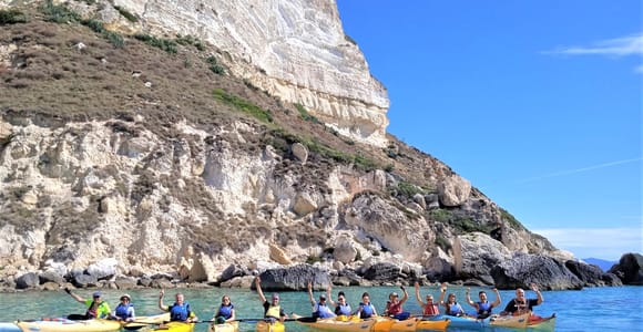 A line of yellow and blue sea kayaks with smiling paddlers in life jackets, waving, below a pale white cliff on turquoise water