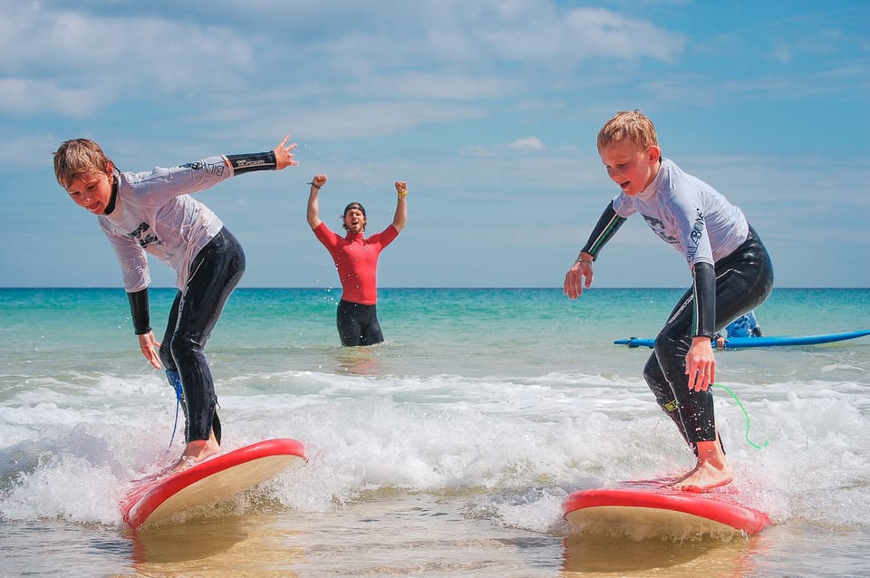 Zwei Kinder surfen kleine Wellen, ein Lehrer steht in der Nähe