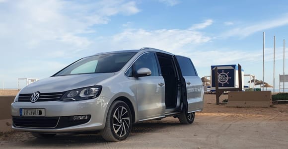 A silver seven-seater van with its sliding door open, parked on packed sand next to a wooden beach bar
