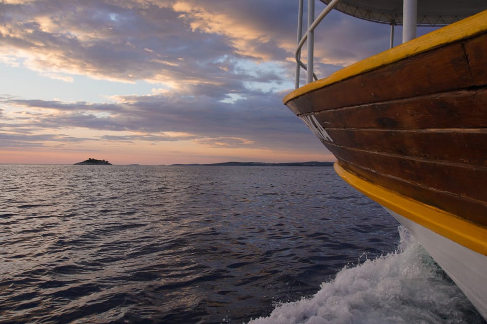 The yellow-railed bow of a wooden boat on calm sea under a pink and grey dusk sky, with a dark island on the horizon