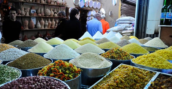Cones of pulses, rice, dried fruit and coloured pasta in metal bowls at a market stall, with earthenware tagine pots and jars stacked on shelves behind
