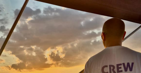 A crew member stands at the bow of a boat looking at the setting sun over the sea