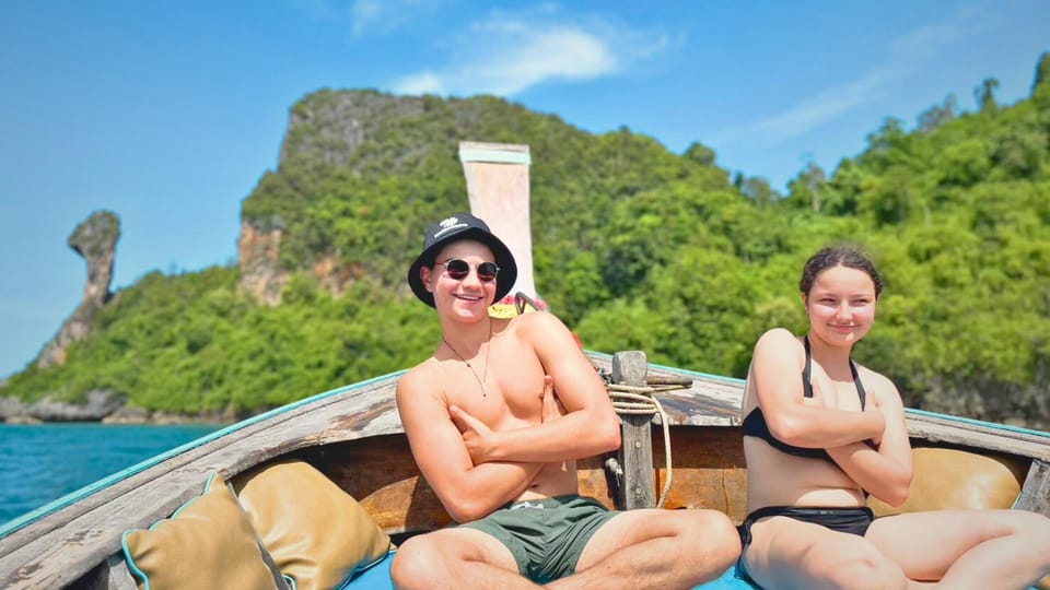 Two travellers sitting on the bow of a longtail boat, the pointed rock of Chicken Island rising behind them