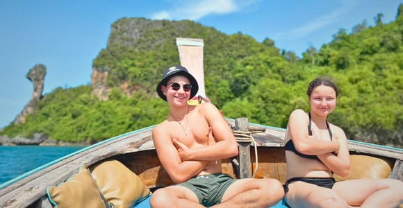Two travellers sitting on the bow of a longtail boat, the pointed rock of Chicken Island rising behind them