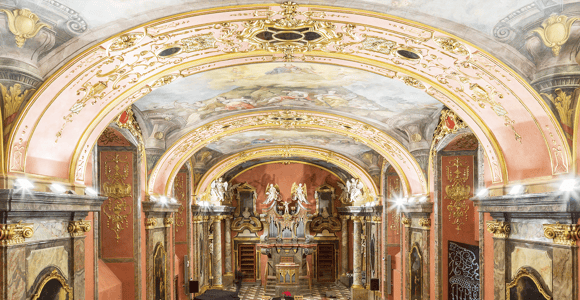 Baroque hall with gilded stucco, a painted vault, an organ at the far end and rows of red chairs