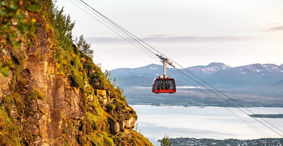 A red cable car gondola on its cable above a green cliff face, with a fjord and a town in the distance