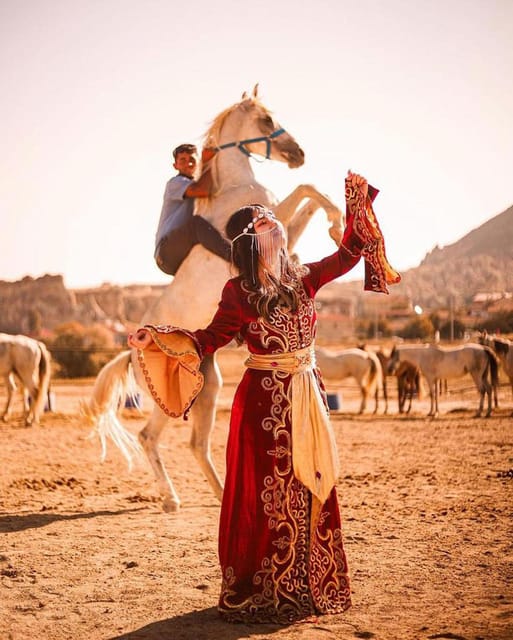 A woman in a red embroidered costume stands in a dusty paddock while a white horse rears behind her