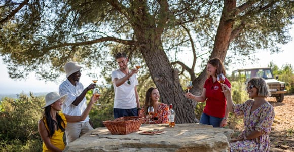 A group toasting with wine at a large stone table under a pine tree, with an off-road vehicle in the background
