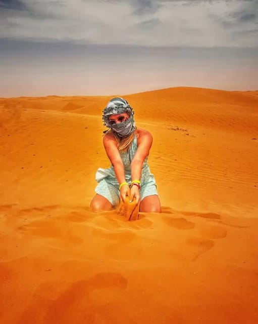 A woman in a checked headscarf sits in orange sand dunes under a grey sky, sand running through her hands
