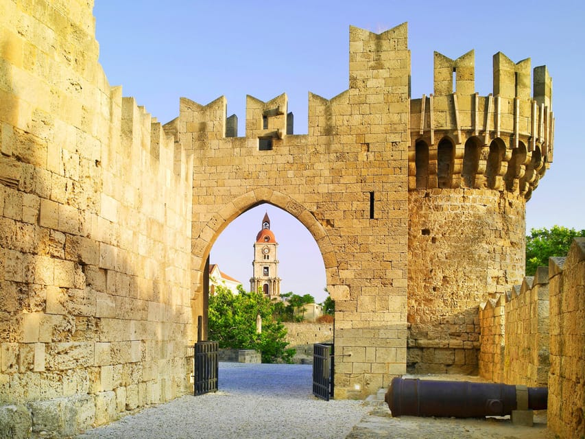 A sandstone fortress gate with a crenellated round tower and a pointed arch, framing a clock tower in the distance under a clear sky