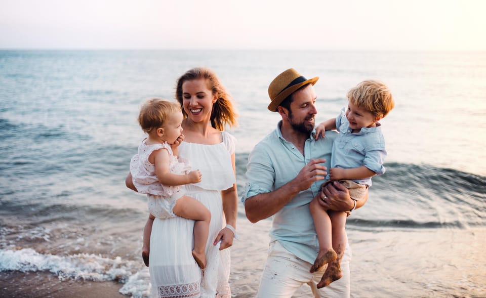 Two parents laughing on a beach, each holding a small child, in soft evening light