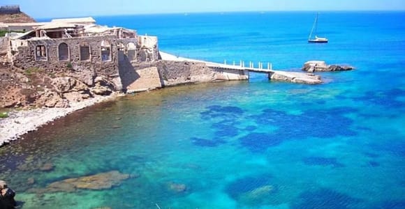 Ruined stone fishing buildings on a rocky shore above clear turquoise water, with a small boat and a distant sailboat