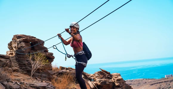 A climber in a harness and helmet on a steel-cabled via ferrata route along a rock face in the dry landscape of southern Gran Canaria