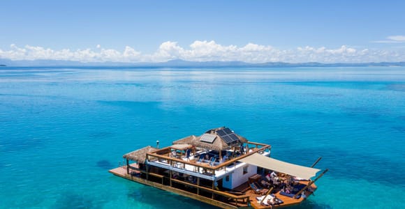 A two-storey wooden pontoon with thatched roofs and solar panels floating alone on turquoise water, guests on both decks under a stretched awning, mountains on the horizon