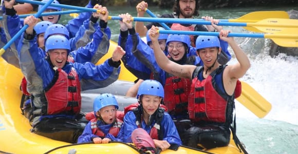 A group in blue helmets and red life jackets raising paddles in a yellow raft on white water
