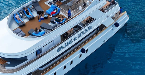 Aerial view of a large white boat named Blue Star, with people lying on cushioned loungers on the open upper deck, on deep blue water
