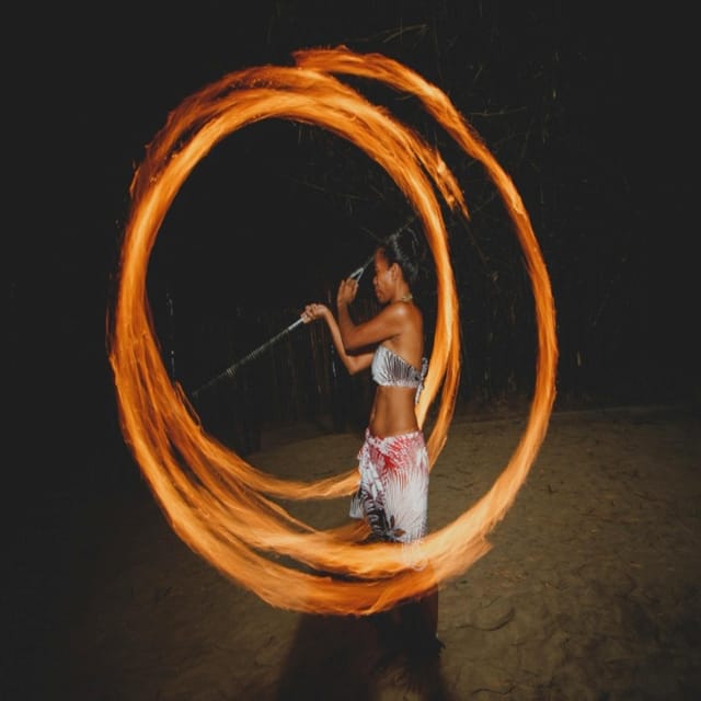 A fire dancer at night drawing a complete circle of flame with a spinning staff, the burning trail lit orange against black, sand underfoot
