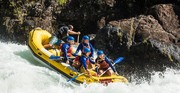 A yellow raft dropping through a wall of white water, five paddlers in blue helmets and red life jackets bracing, a guide steering at the stern beside a dark rock face