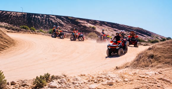 Several riders on red quad bikes with helmets on a dusty sandy track curving round a dry hillside under a clear blue sky