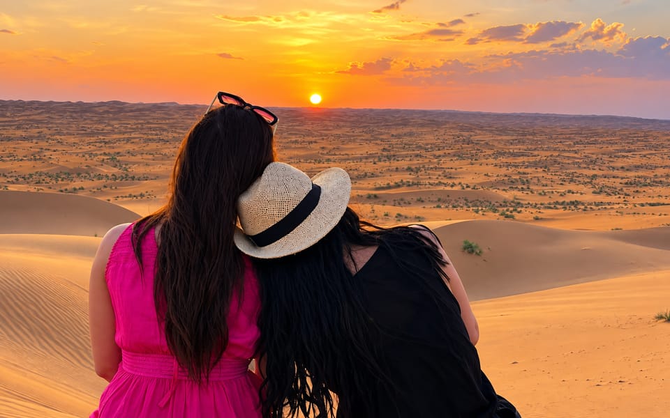 Two women sit close together on a dune crest, seen from behind, watching a low orange sun set over ridge after ridge of sand under a pink and gold sky