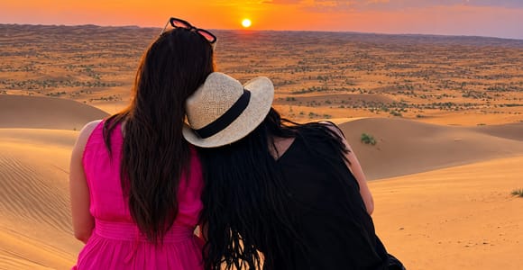 Two women sit close together on a dune crest, seen from behind, watching a low orange sun set over ridge after ridge of sand under a pink and gold sky