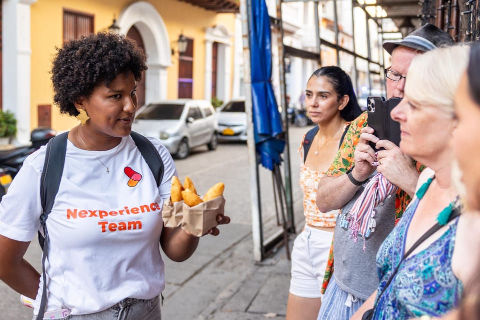 A guide in a white T-shirt holding out a paper bag of golden fried pastries to a small group on a pavement, one guest filming with a phone, a yellow colonial façade behind