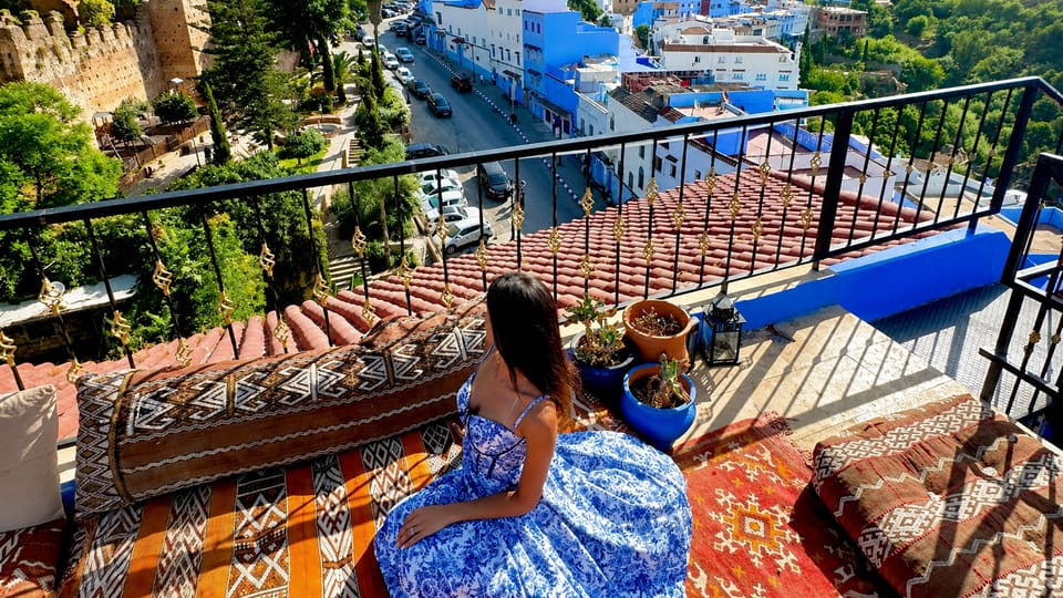 A woman in a blue patterned dress sits on cushions on a rooftop terrace above blue-washed houses and a street lined with parked cars