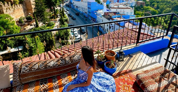 A woman in a blue patterned dress sits on cushions on a rooftop terrace above blue-washed houses and a street lined with parked cars