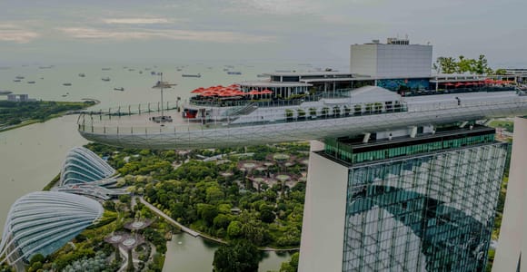 The cantilevered roof terrace of Marina Bay Sands seen from above, with the domes and Supertrees of the Gardens on the ground below and ships anchored offshore