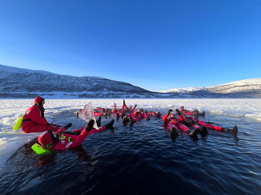 A group of people in red survival suits floating on their backs in dark water among ice floes, with snowy mountains behind
