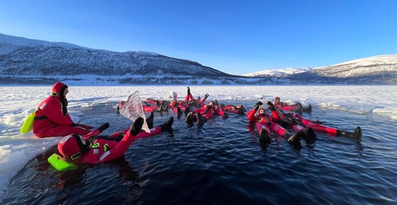 A group of people in red survival suits floating on their backs in dark water among ice floes, with snowy mountains behind