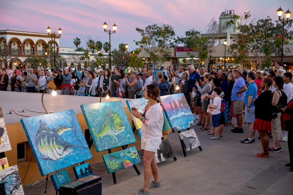 A crowd on a town square at dusk watching a painter beside easels of bright fish paintings, colonial arcades and a church tower behind
