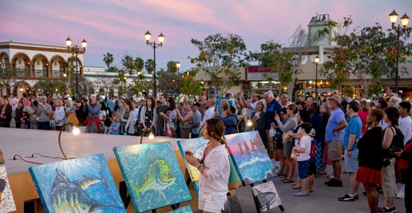A crowd on a town square at dusk watching a painter beside easels of bright fish paintings, colonial arcades and a church tower behind