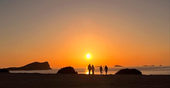 Four people in silhouette walking across a beach toward a low orange sun, with rocky islets on the horizon