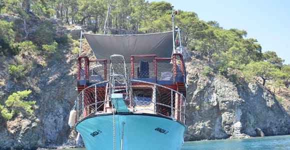 Turquoise wooden gulet anchored in a calm bay below a rocky, pine-covered shore