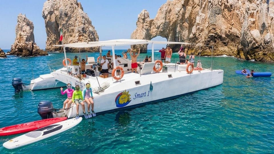 A white catamaran full of passengers on turquoise water in front of the Land's End rocks, three children in snorkel masks sitting on the stern