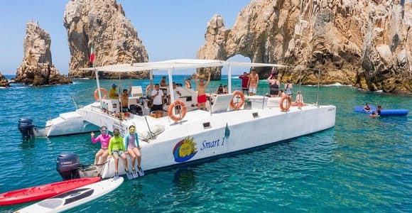 A white catamaran full of passengers on turquoise water in front of the Land's End rocks, three children in snorkel masks sitting on the stern