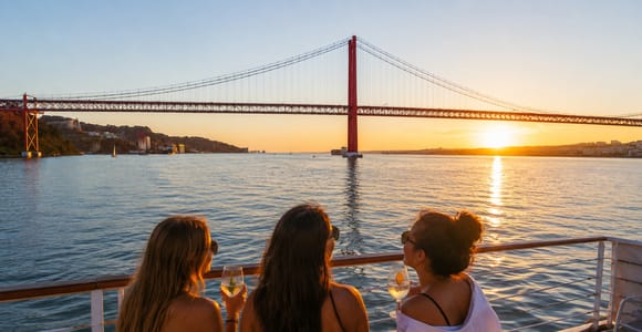 Three people in summer clothes on the open rear deck of a boat, wine glasses in hand, the red steel of a suspension bridge and a low sun over the water behind them