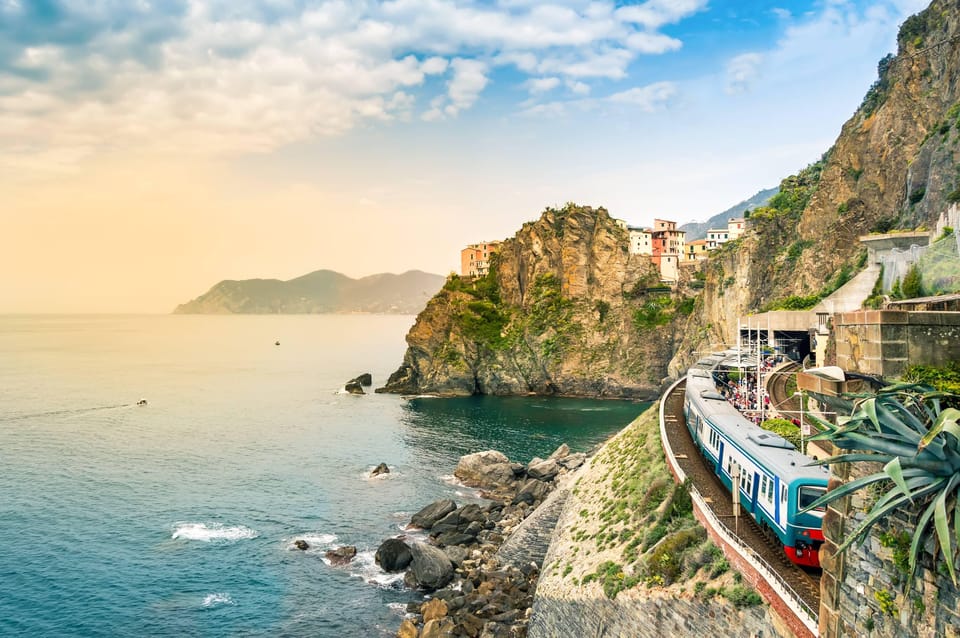 Blue and white regional train running along a rocky cliff above the sea, with a village perched on the headland ahead