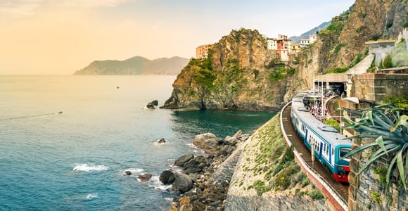 Blue and white regional train running along a rocky cliff above the sea, with a village perched on the headland ahead