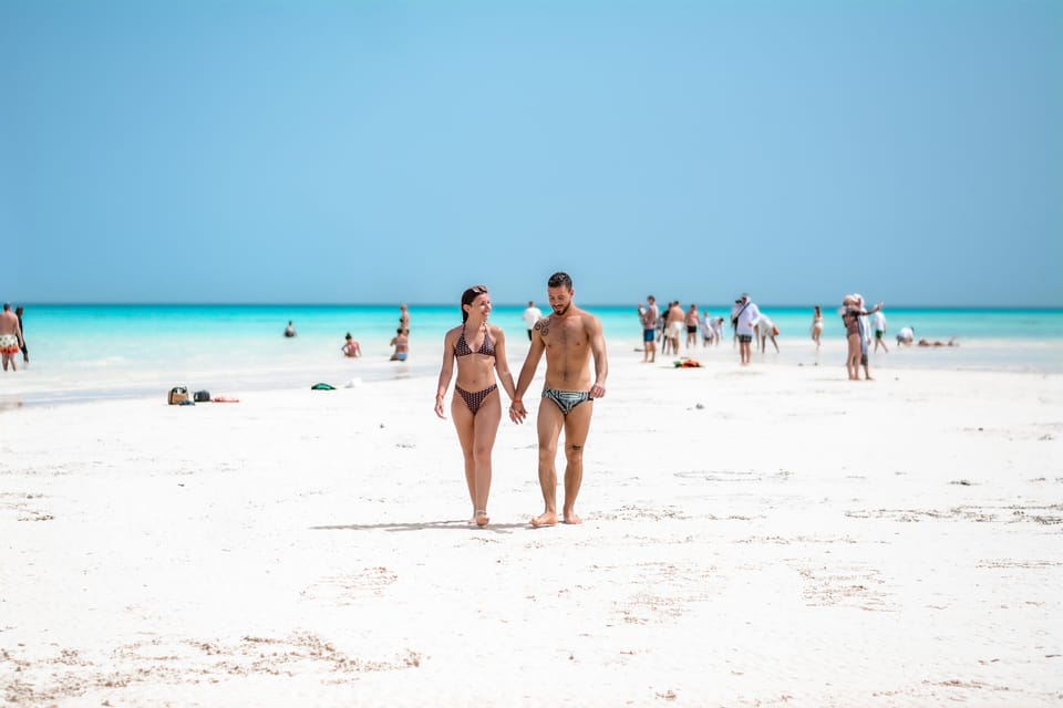 Una coppia cammina a piedi nudi lungo un’ampia lingua di sabbia bianca, con acque basse turchesi su entrambi i lati e altri visitatori in piedi nell’acqua sullo sfondo