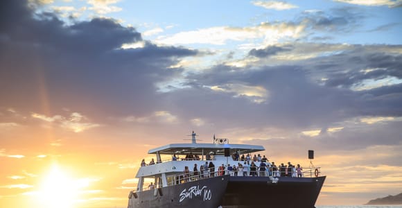 A dark double-deck catamaran crowded with passengers on the upper deck, the low sun beside it laying a bright track across the water