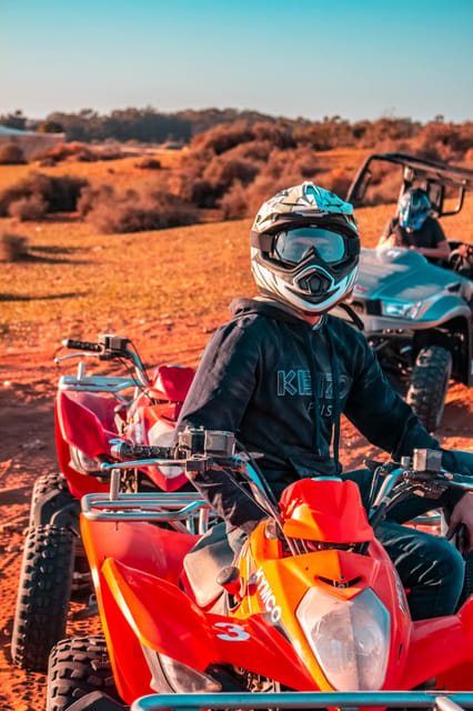 A rider in a white helmet and goggles sits on a red quad bike on red earth, with a second quad behind