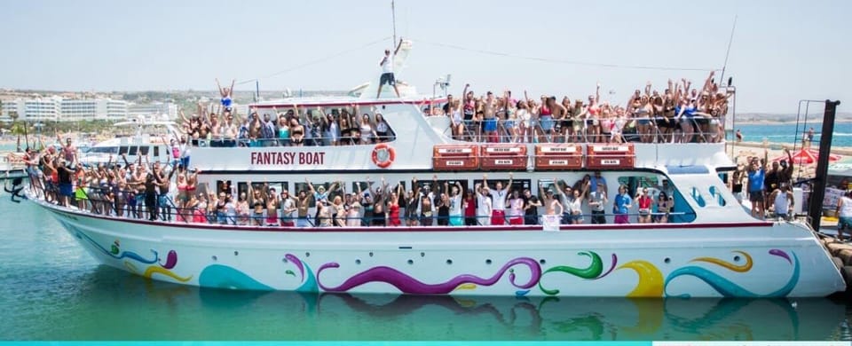 Crowd of people waving from the decks of a large white party boat named Fantasy Boat, moored in a harbour in bright sunshine