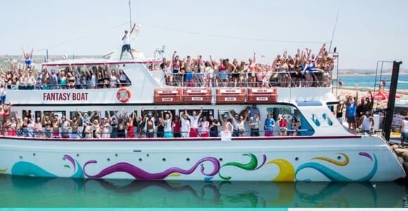 Crowd of people waving from the decks of a large white party boat named Fantasy Boat, moored in a harbour in bright sunshine
