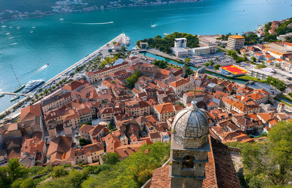 Terracotta roofs of Kotor’s old town seen from above, with a church dome and the bay at left