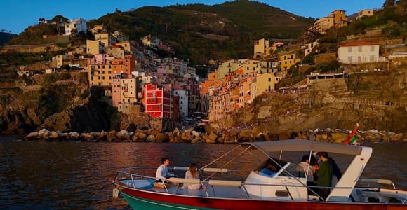 Small motorboat with an awning on dark water in front of Riomaggiore's tall orange and pink houses glowing in evening light