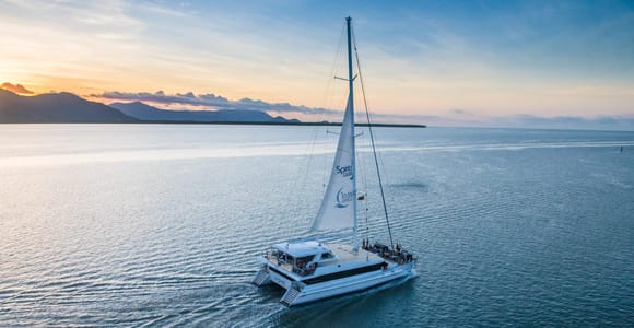 A white sailing catamaran with its mainsail up, photographed from above on flat grey water at dusk, a low dark headland and an orange sky behind it