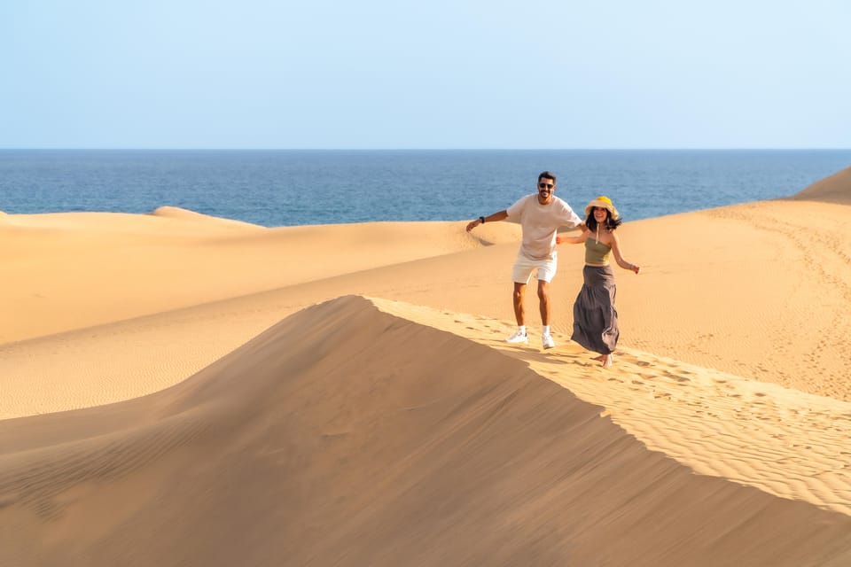 A couple walking across rippled Maspalomas sand while a photographer frames them in warm evening light